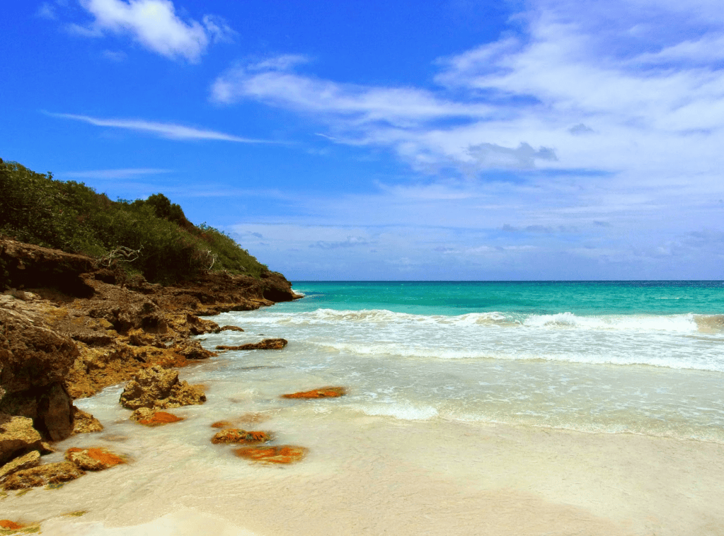 Sunny beach with rocky shoreline, clear blue sky, and gentle waves.