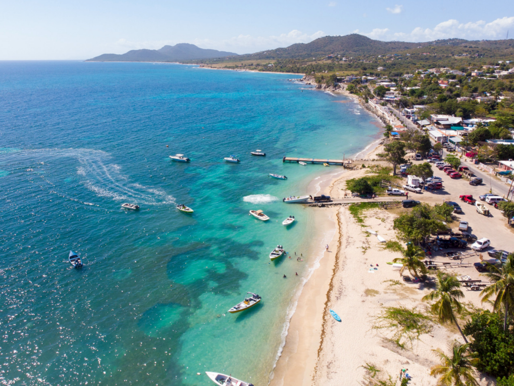 Aerial view of a coastal beach with boats on turquoise water and a shoreline with trees and buildings.