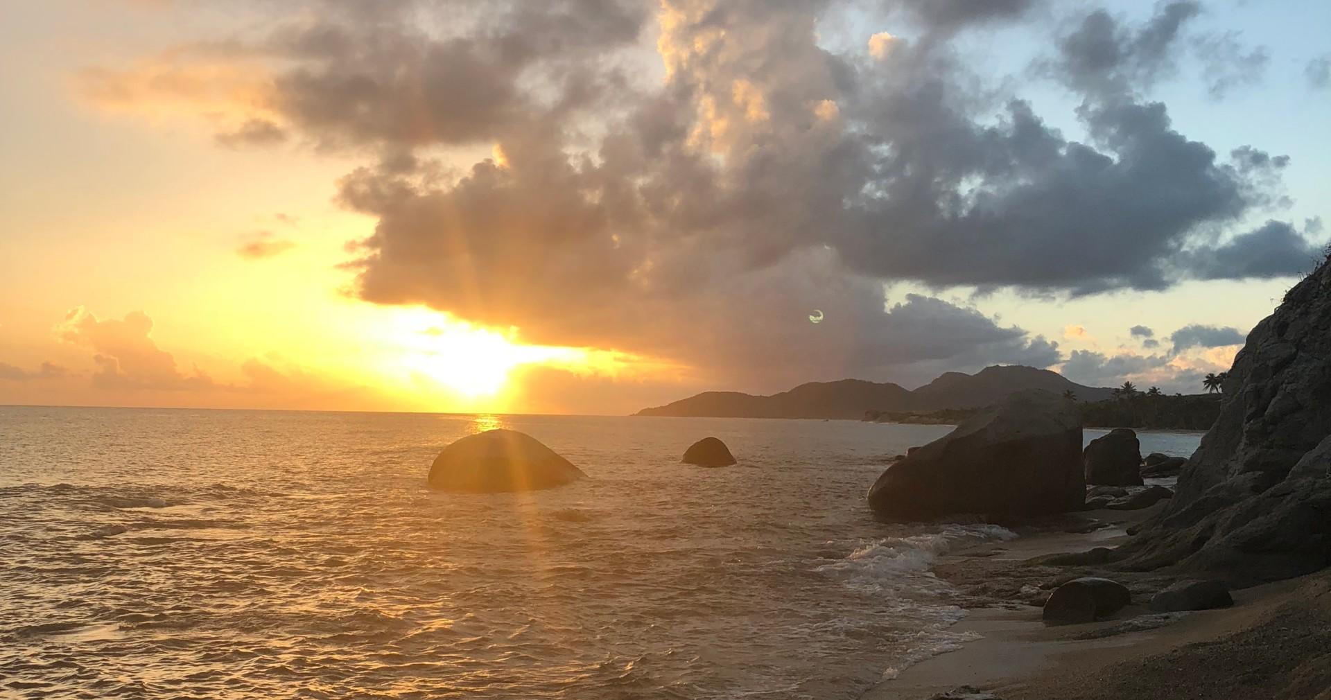 Sunset over ocean with rocky shoreline and clouds in sky.